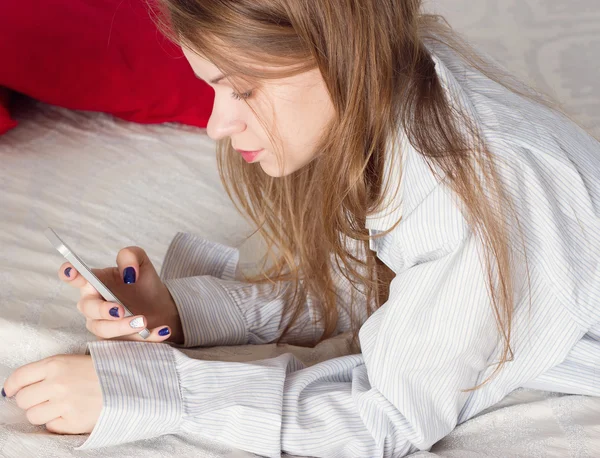 Дівчина лежить у чоловічій сорочці на ліжку з смартфоном — стокове фото