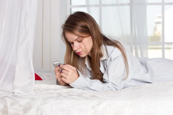 Girl lies in a mans shirt on the bed with smartphone — Stok fotoğraf