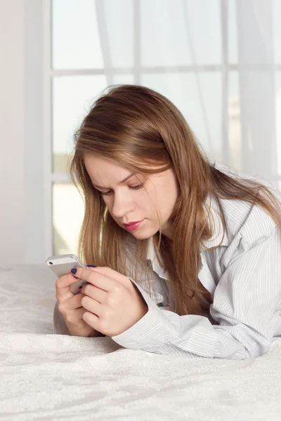 Girl lies in a mans shirt on the bed with smartphone — Stock fotografie