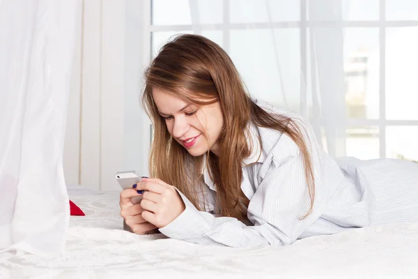 Girl lies in a mans shirt on the bed with smartphone — Stok fotoğraf