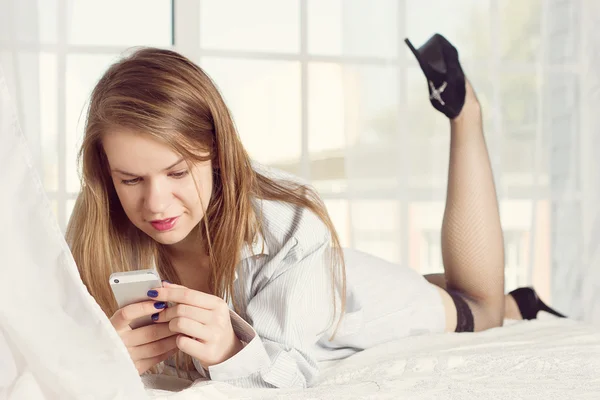 Girl lies in a mans shirt on the bed with smartphone — Stock fotografie