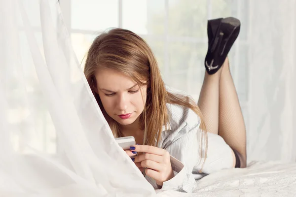 Menina encontra-se em uma camisa de homem na cama com smartphone — Fotografia de Stock