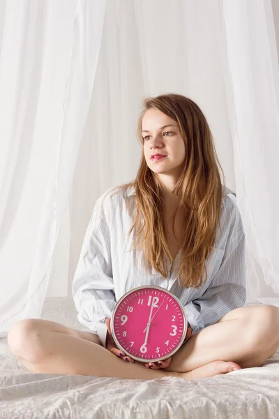 Chica está sentada en la cama con un gran reloj rosa. 6 a.m. —  Fotos de Stock