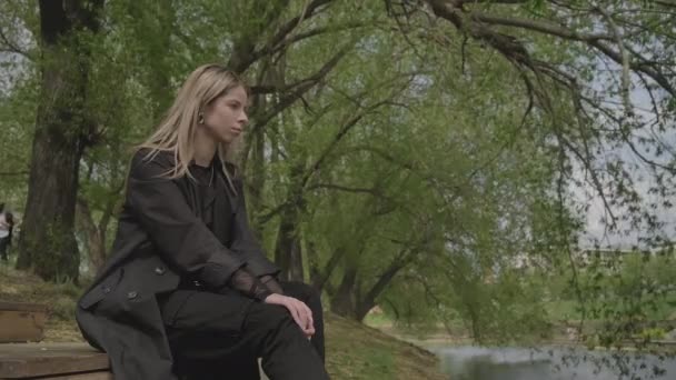 En verano, una chica se sienta en un muelle de madera cerca del agua, mira a la distancia y está triste — Vídeos de Stock