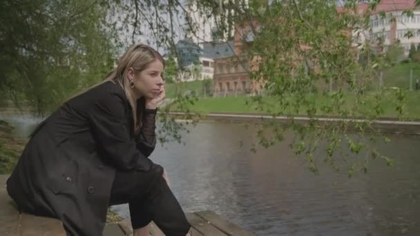 En verano, una chica se sienta en un muelle de madera cerca del agua, mira a la distancia y está triste — Vídeos de Stock