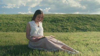 A girl sits on the grass, reads a book and looks calmly into the distance