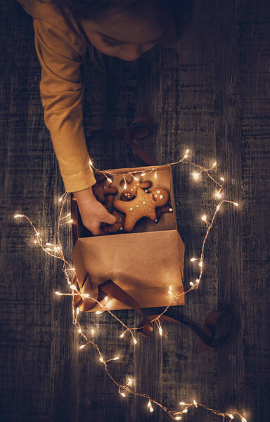 Child Received Gift Box with Christmas Cookies Decorated with Glowing Garland. Little Baby Eating Gingerbread. Happy Winter Holidays.