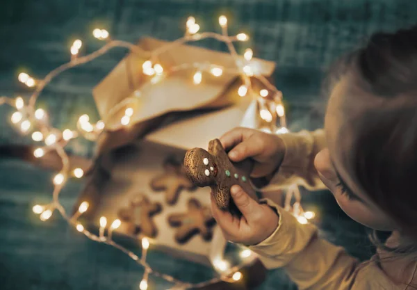 孩子收到的礼物盒与圣诞饼干装饰与发光的加兰 小婴儿吃姜饼 寒假快乐 — 图库照片