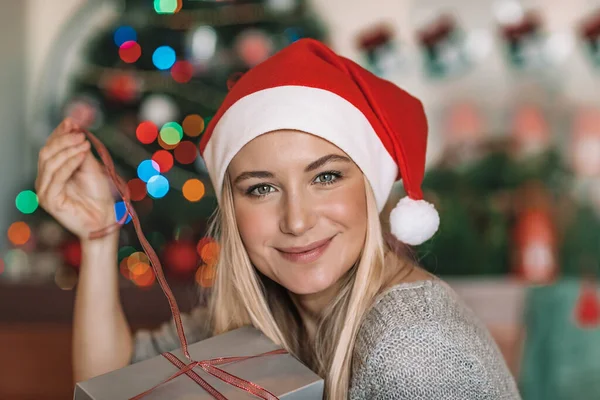 一个快乐的金发女人穿着红色圣诞老人帽 带着礼品盒站在圣诞树旁的画像 快乐的圣诞老人助手 温馨的平安夜在家里 — 图库照片