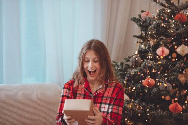 Heyecanlı Genç Kadın Noel Hediyesini Açıyor, Dekorasyon Ağacı 'nın yanında oturuyor, Kırmızı ekose pijama giyiyor, Hoş İfade Ediyor, Elleri Tutan Hediye