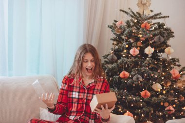Heyecanlı Genç Kadın Noel Hediyesini Açıyor, Dekorasyon Ağacı 'nın yanında oturuyor, Kırmızı ekose pijama giyiyor, Hoş İfade Ediyor, Elleri Tutan Hediye