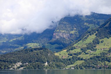 Tepenin üstünde bulutlar var. Çimenli ve ağaçlık alanları görebilirsiniz.