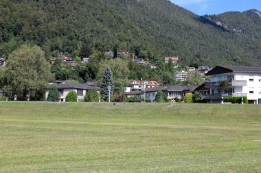 Uzakta, çimenli arazinin arkasında yamacında ağaçlık alanlar olan bir dağ tepesi görülür. Bu İsviçre 'deki Schwyz kantonunda Brunnen' de görülür..