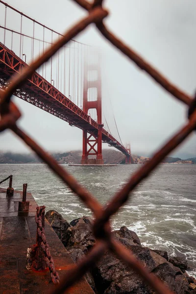 The famous Golden Gate bridge viewed through a rusty chain link fence on a cloudy day in San Francisco, California — Stock Photo, Image
