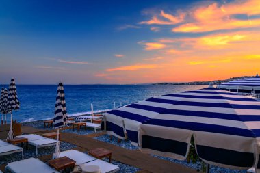 Mavi ve beyaz çizgili şemsiyeler, Akdeniz boyunca uzanan altın bir günbatımında Fransa 'nın başkenti Anglais' te bulunan gezinti güvertesi boyunca çakıl taşlı bir plaj hizasında.