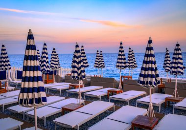 Mavi ve beyaz çizgili şemsiyeler, Akdeniz boyunca uzanan altın bir günbatımında Fransa 'nın başkenti Anglais' te bulunan gezinti güvertesi boyunca çakıl taşlı bir plaj hizasında.