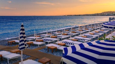 Mavi ve beyaz çizgili şemsiyeler, Akdeniz boyunca uzanan altın bir günbatımında Fransa 'nın başkenti Anglais' te bulunan gezinti güvertesi boyunca çakıl taşlı bir plaj hizasında.