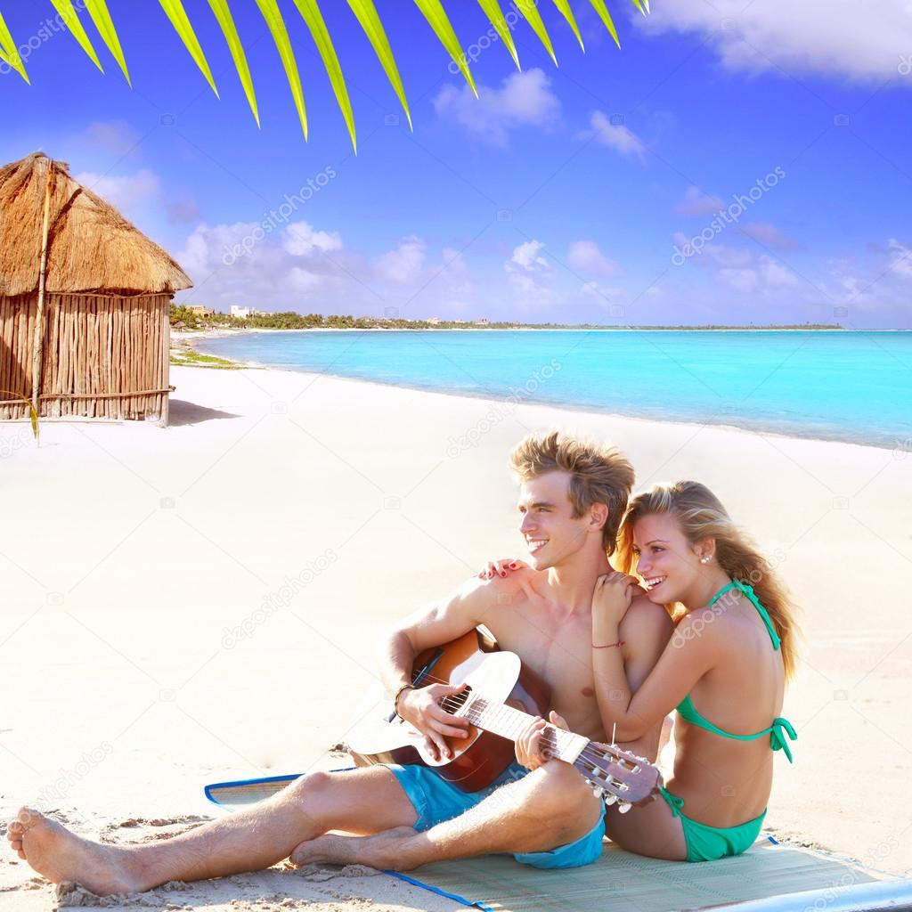 Blond couple sitting in a beach sand playing guitar