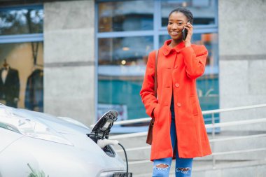 Beklerken akıllı telefon kullanmak. Elektrikli araba şarj istasyonundaki Afrikalı Amerikalı kız. Yepyeni bir araç..