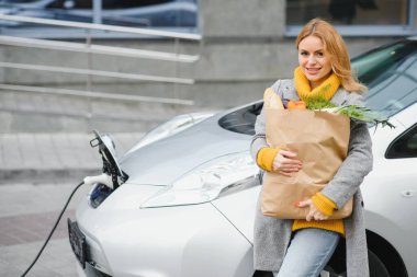 Elektrikli benzin istasyonunda elektrikli araba şarj ediliyor. Arabanın yanında duran kadın. Yemekleri olan kadın..