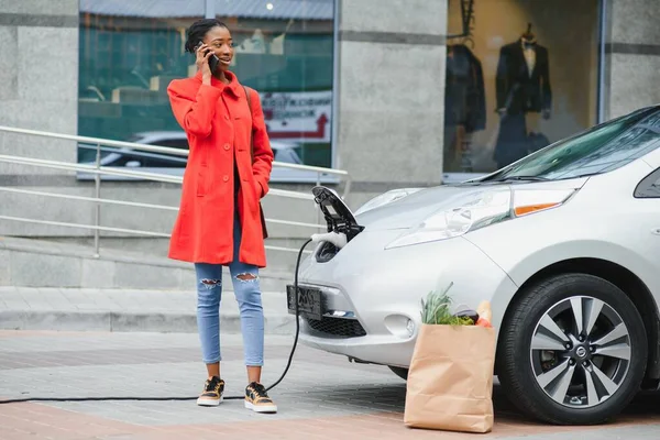 Charging electro car at the electric gas station. Woman standing by the car. Lady with foodstuff.