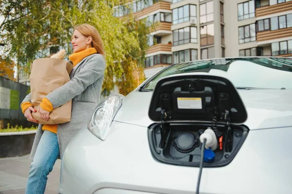 Charging electro car at the electric gas station. Woman standing by the car.