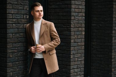 İş adamı elinde kahveyle toplu taşıma binasının önünde yürüyor ve başka tarafa bakıyor. İş gezisindeki yakışıklı genç adam..