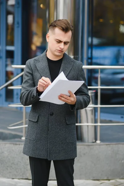 Portrait of serious businessman looking papers on city street. Worried manager reading documents in stylish suit outside. Disappointed executive working with documents outdoors