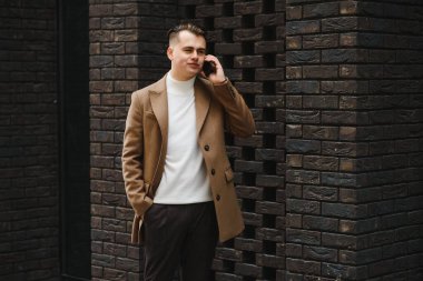 Yakışıklı, gülümseyen genç işadamı. Parmaklıklara yaslanmış bir şekilde. Şehir caddesinde duruyor, elinde cep telefonu var.