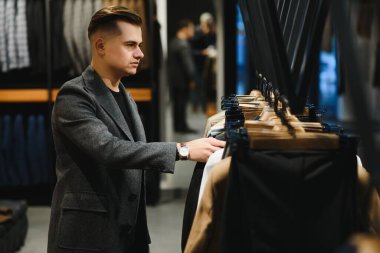 Bir kostüm dükkanındaki genç ve yakışıklı işadamı askıdaki ceketin kolundaki kumaşı inceliyor..