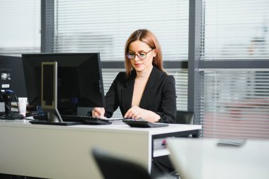 Güzel, hoş, sevimli, mükemmel kadın çalışma masasında deri sandalyede oturuyor, gözlük takıyor, resmi kıyafetler giyiyor, dizüstü bilgisayarı ve defteri masada.