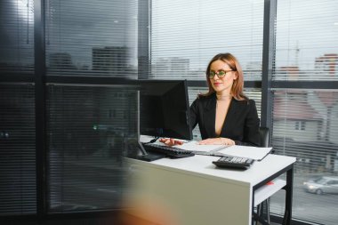 Güzel, hoş, sevimli, mükemmel kadın çalışma masasında deri sandalyede oturuyor, gözlük takıyor, resmi kıyafetler giyiyor, dizüstü bilgisayarı ve defteri masada.