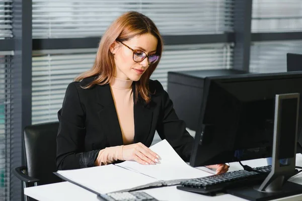 Pretty, nice, cute, perfect woman sitting at her desk on leather chair in work station, wearing glasses, formalwear, having laptop and notebook on the table