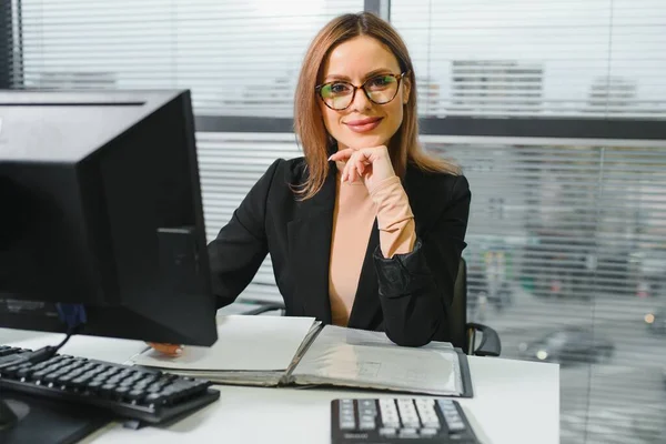 Pretty, nice, cute, perfect woman sitting at her desk on leather chair in work station, wearing glasses, formalwear, having laptop and notebook on the table