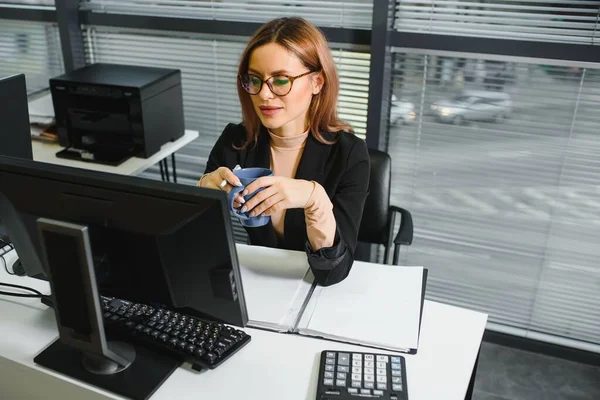 Pretty, nice, cute, perfect woman sitting at her desk on leather chair in work station, wearing glasses, formalwear, having laptop and notebook on the table