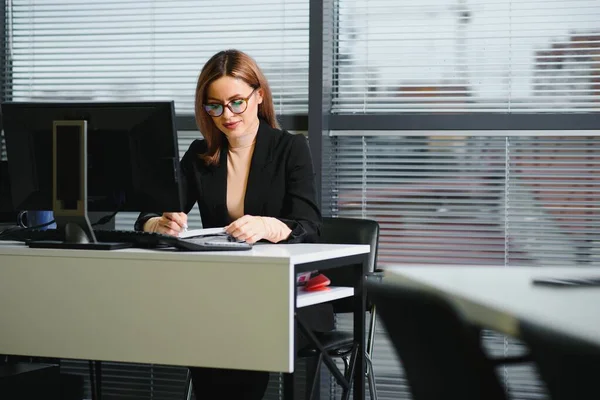 Pretty, nice, cute, perfect woman sitting at her desk on leather chair in work station, wearing glasses, formalwear, having laptop and notebook on the table