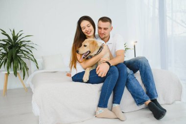 Neşeli esmer genç bayan en sevdiği köpeğiyle yatakta oynarken kocası dizüstü bilgisayarında sosyal ağlarda geziniyor hafta sonlarını yatak odasında geçiriyor. İç atmosfer