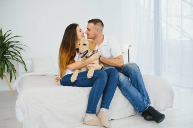 Neşeli evli çift evde köpekle oynuyor. Kanepede oturup hayvanı okşuyorlar. Kadın ve erkek gülümsüyor.