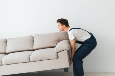 Yükleyici kanepeyi, kanepeyi taşıyor. Tulumlu işçi kanepeyi kaldırıyor, beyaz arka plan. Teslimat hizmeti konsepti. Kurye taşınma ya da taşınma ihtimaline karşı mobilya taşır..
