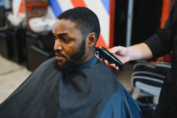 Berber dükkanında profesyonel elektrikli makasla kırpılan genç siyahi bir adamın portresi. Erkek güzellik tedavi konsepti. Genç Afrikalı adam berber salonunda yeni saç kesimi yaptırıyor.