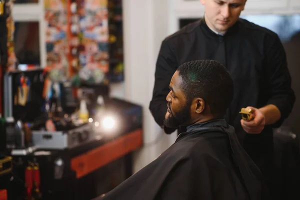 Portrait of young black man being trimmed with professional electric clipper machine in barbershop.Male beauty treatment concept. Young African guy getting new haircut in barber salon