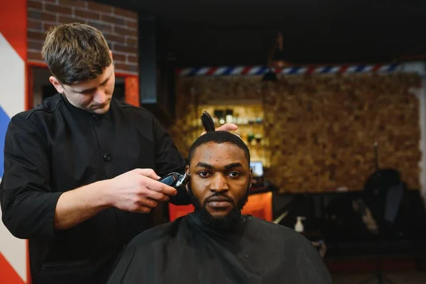Portrait of young black man being trimmed with professional electric clipper machine in barbershop.Male beauty treatment concept. Young African guy getting new haircut in barber salon