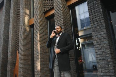 İş iletişimi. Resmi kıyafetli mutlu bir Afrikalı adam cep telefonuyla konuşuyor ve dışarıda dururken gülümsüyor.