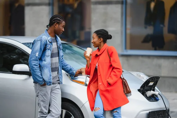 Happy young adult african american man and smiling woman charging electric car.