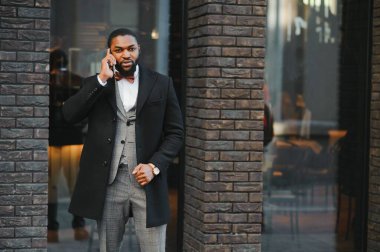 İş iletişimi. Resmi kıyafetli mutlu bir Afrikalı adam cep telefonuyla konuşuyor ve dışarıda dururken gülümsüyor.