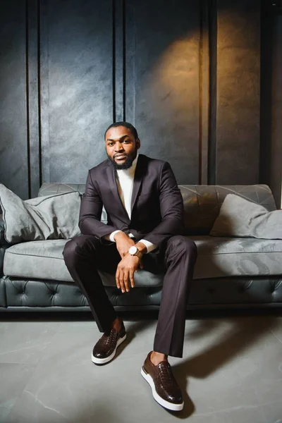 Young black man in suit on dark background sitting on a sofa. Fashion portrait of young man.