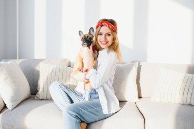 Neşeli genç bir kadın siyah burunlu köpeğini tutuyor ve gülüyor. Fransız buldoguyla poz veren gülümseyen kızın kapalı bir portresi.