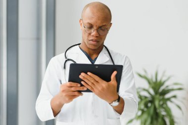 Koridorda dijital tablet kullanan olgun bir Afrikalı doktor. Klinikte fotokopi alanı olan tablet bilgisayar kullanan kendine güvenen erkek doktorun portresi. Steteskop takan başarılı gülümseyen doktor..