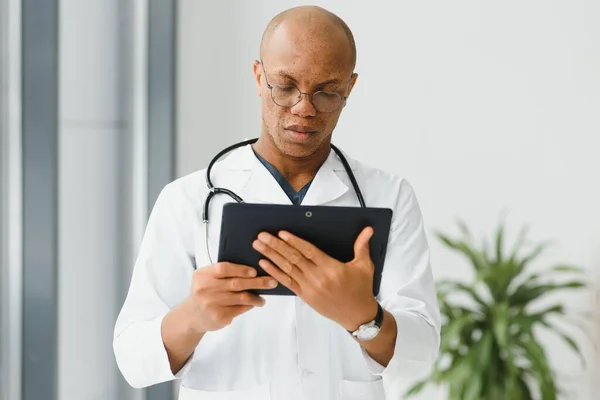 Mature african doctor using digital tablet in corridor . Portrait of confident male doctor using tablet computer in clinic with copy space. Successful smiling doctor in labcoat wearing stethoscope.