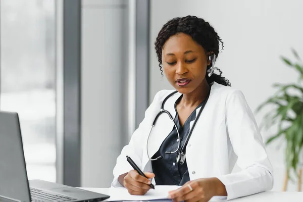 medicine, online service and healthcare concept - happy smiling african american female doctor or nurse with headset and laptop having conference or video call at hospital